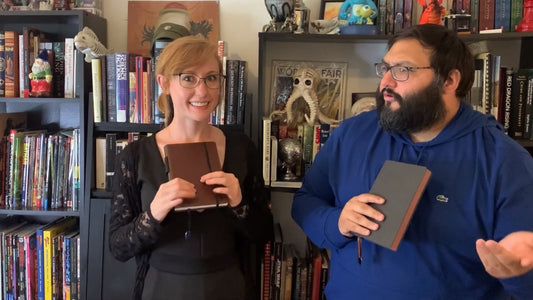 Two reviewers hold brown and black LeStallion notebooks in front of bookshelves during an unboxing video.