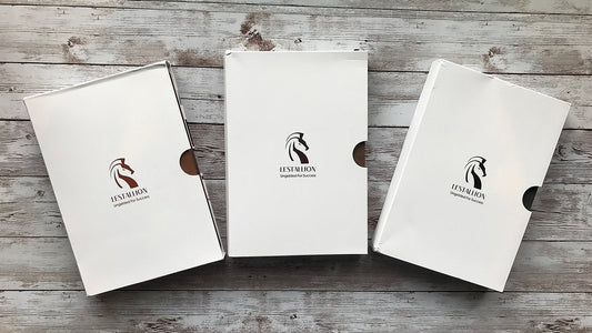 Three LeStallion notebooks—Henson Desert Brown, Garanno Cocoa Brown, and Ariegeois Ashgray Black—on gray faux fur beside white flowers.